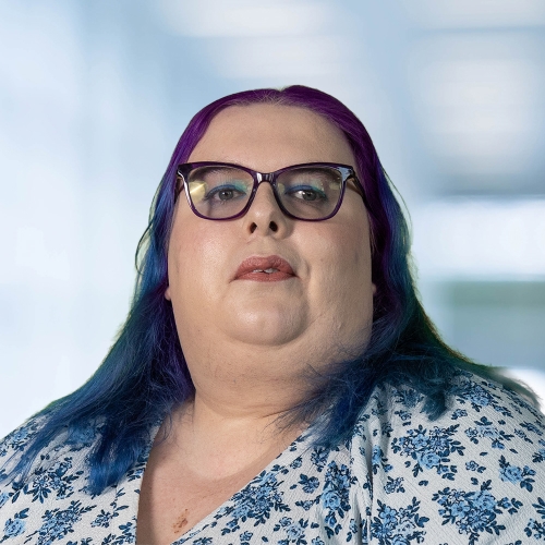 A photo of Emma Atkins. She has coloured hair in shades of dark blue and purple and is wearing glasses. She wears a floral white top.