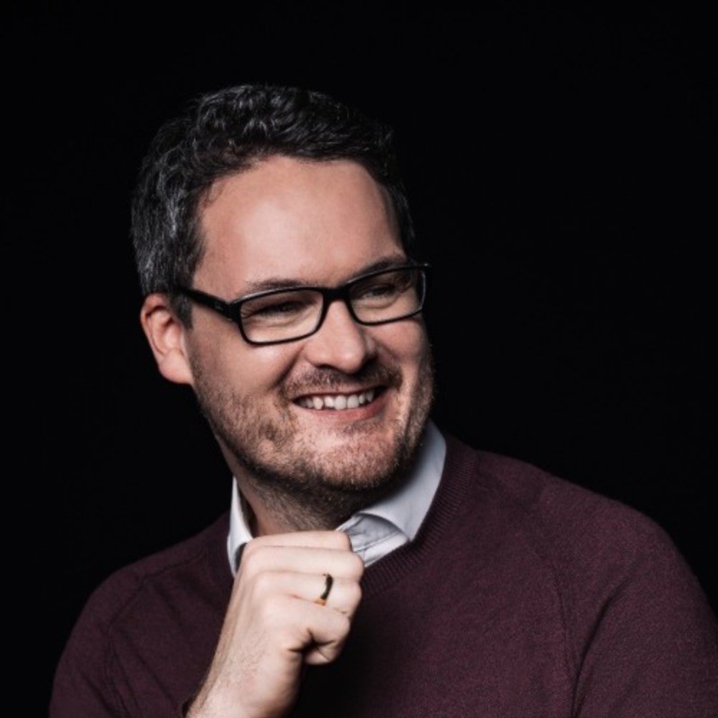 A photo of Timo Graf von Koenigsmarck smiling. He has short dark hair and a beard. He wears dark glasses and a red jumper.