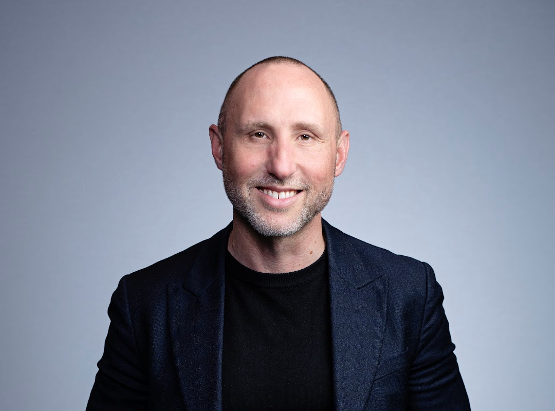 A bald man dressed in a black shirt and jacket poses, exuding a polished and sophisticated look.