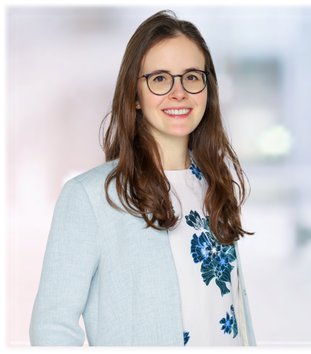 A photo of Hortense Fricker smiling. She has just below shoulder hair and wears dark round glasses and a floral top with dark blue flowers and a sky blue blazer.