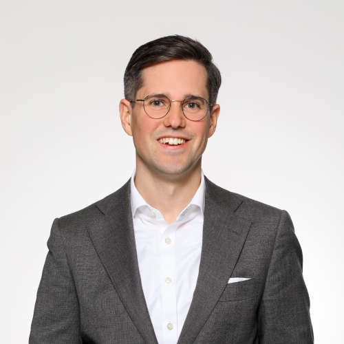 A photo of Julian Brettschneider Lázaro smiling. He has short dark hair. He wears dark glasses like Harry Potter and a white shirt with grey blazer.