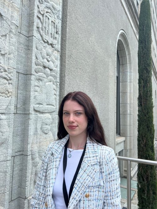 A photo of Lena Stoffer. She has brownish long hair. She wears white top and a lining blazer and an id around the neck.