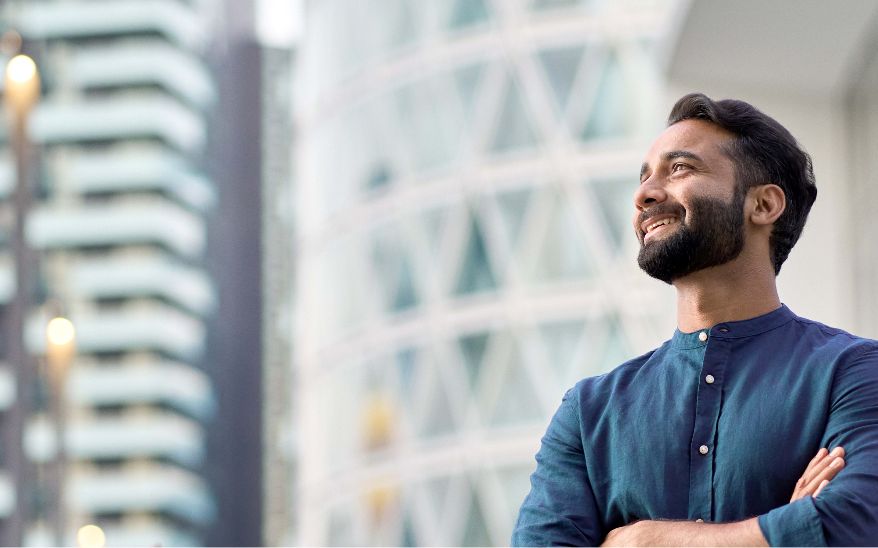 Person with blurred face in blue shirt standing with arms crossed in front of modern high-rise buildings.