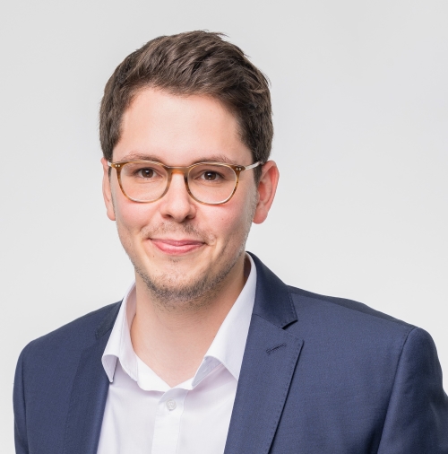 A photo of Malte Jünger smiling. He has short dark hair and a stubble. He wears light framed glasses and a white shirt with steel grey blazer