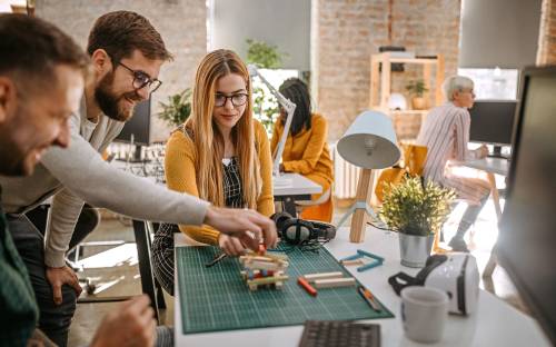 Een team van twee mannen en een vrouw bouwt enthousiast met rechthoekige blokjes, terwijl op de achtergrond collega's druk aan het werk zijn.