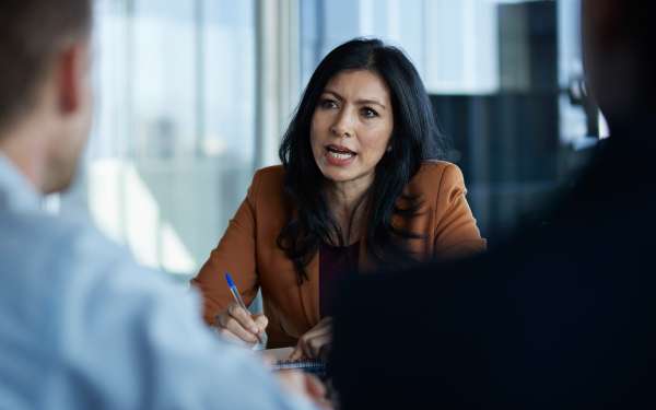 Person in a brown blazer holding a pen during a meeting in a modern office setting.