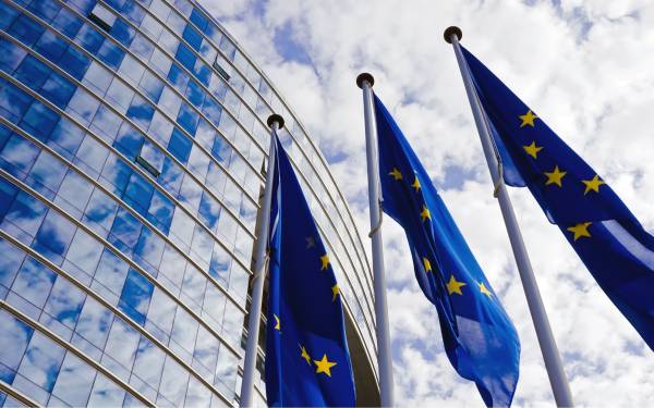 Three European Union flags in front of a modern glass building with a partly cloudy sky.