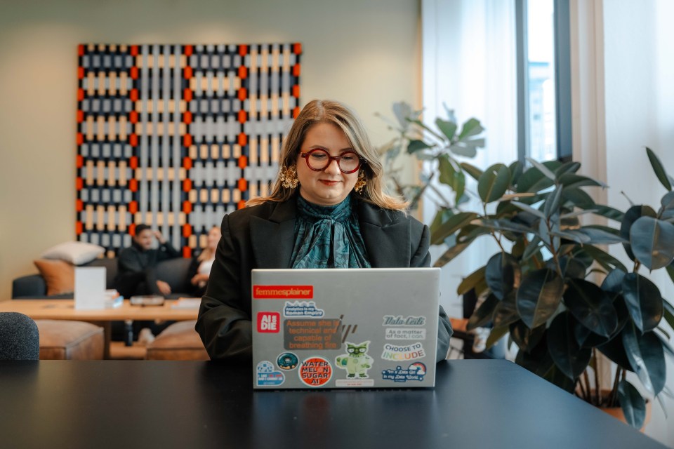Vitalija Bartuseviciute sitting at a desk with a sticker-covered laptop in an office setting, with plants and patterned wall decor.