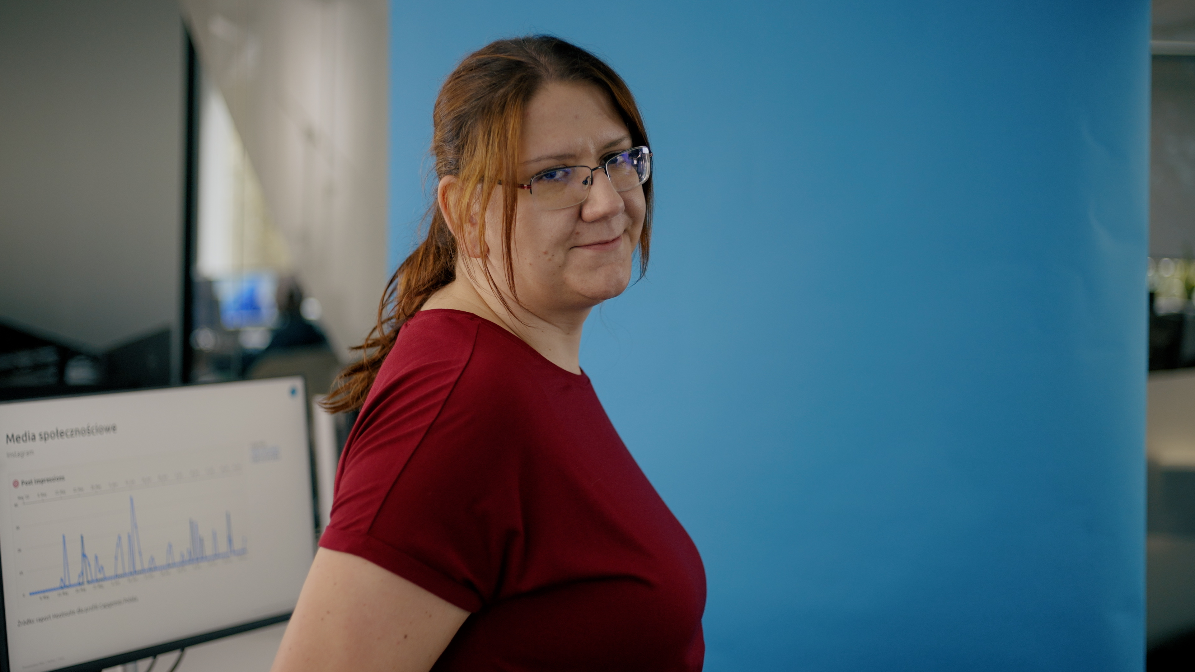 Every Desk is a story - Ania Bąk na niebieskim tle, stojąca bokiem i uśmiechająca się do kamery