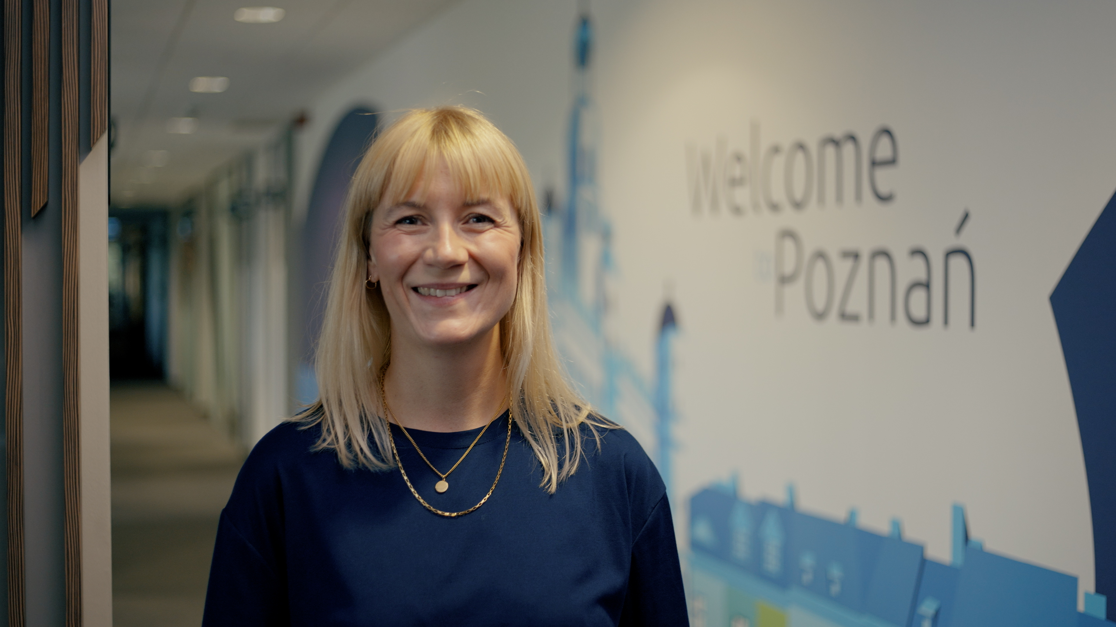 Every Desk is a story - Barbara Górnikiewicz na tle biura Capgemini w Poznaniu w granatowej sukience uśmiechająca siędo aparatu