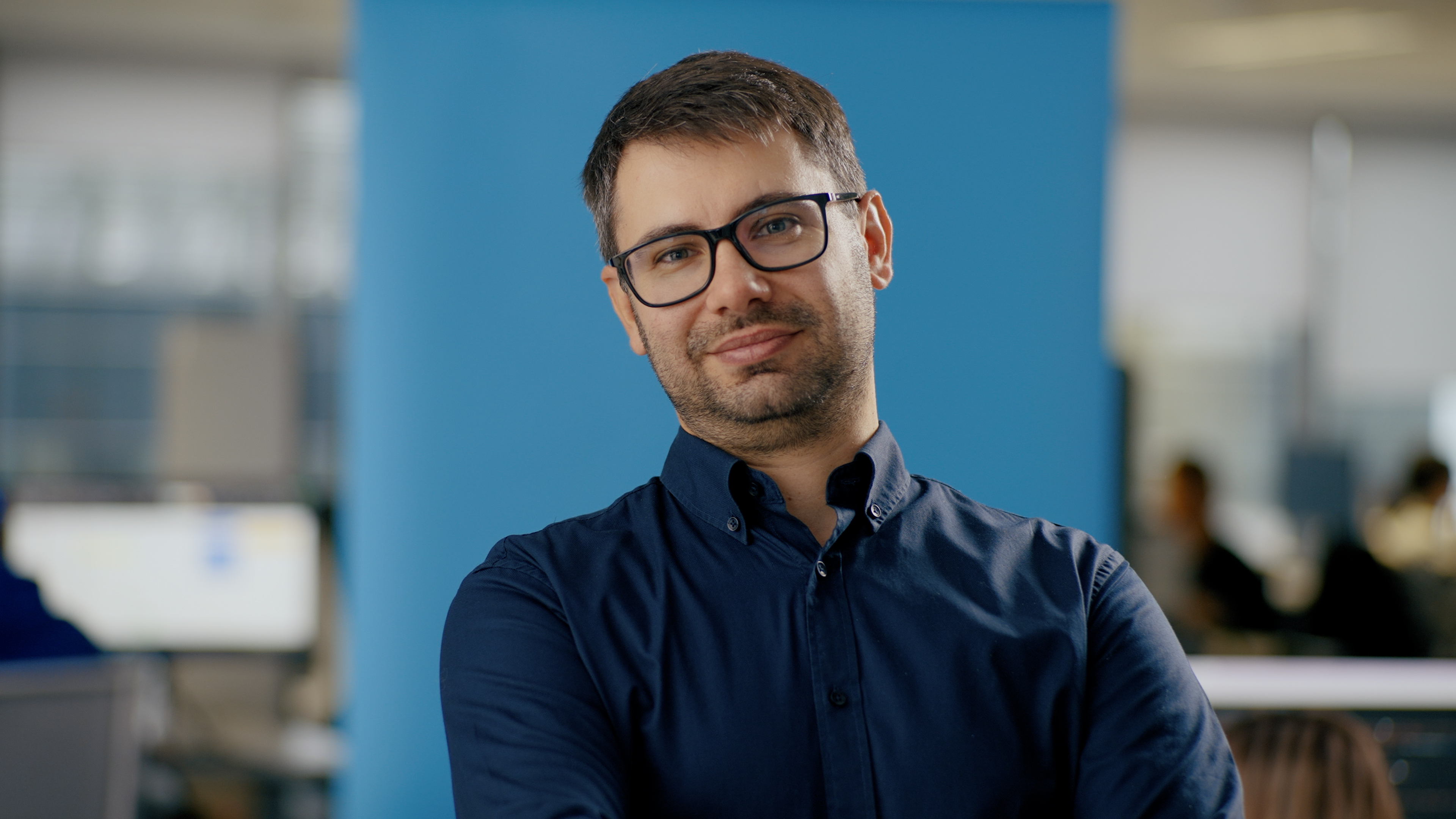 Every Desk is a Story - Vassil Stoitsev na niebieskim tle w granatowej koszuli w okularach uśmiechający się do kamery