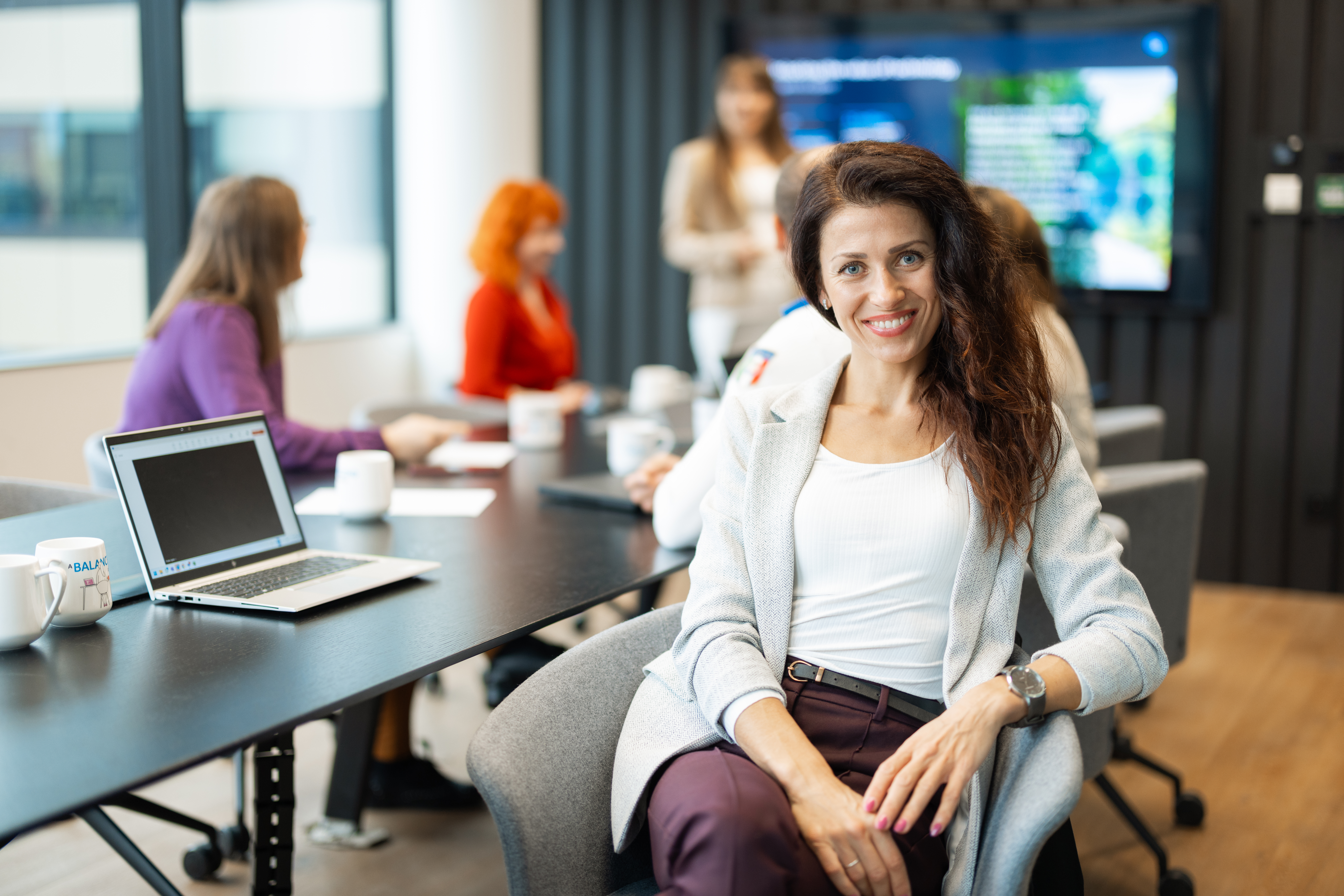 a young lady is smiling to the camera, behind her a group of people in the conference room