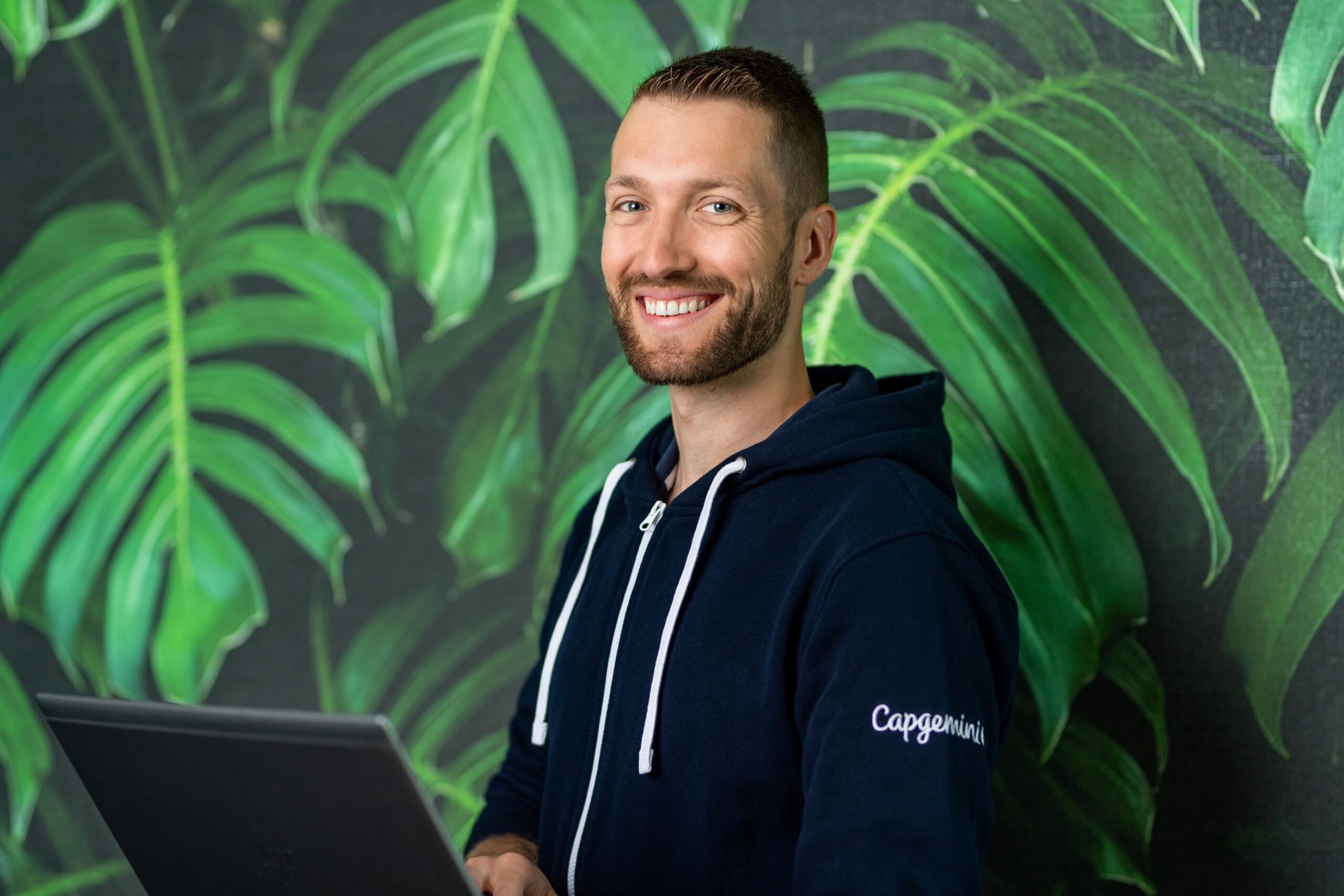 Grzegorz Paprzycki w granatowej bluzie z logo Capgemini trzymający laptopa na tle fototapety w liście