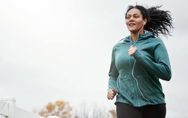 Person running outdoors in a green hoodie and black pants, with autumn trees and a cloudy sky in the background.
